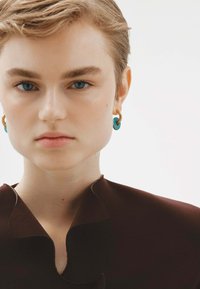 Prenda marrón con un escote festoneado; pendientes azules semi-translúcidos con aros dorados. Textura suave, diseño minimalista.