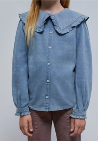 Camicia in denim azzurro chiaro con un ampio colletto arricciato, chiusura con bottoni e delicati ricami floreali. Maniche lunghe con polsi arricciati.