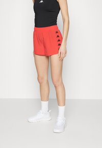 Pantalones cortos deportivos rojos con cintura elástica, con logotipos laterales en negro. Combinados con una camiseta negra, zapatillas blancas y calcetines blancos.
