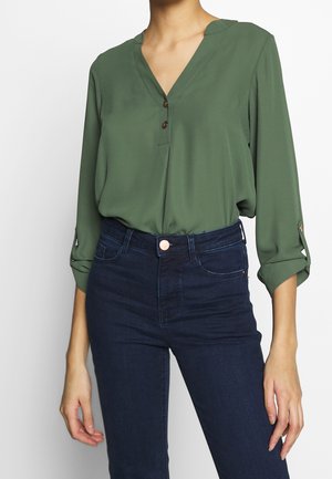 Femme portant un chemisier vert à col en V avec des manches retroussées et un jean taille haute bleu foncé, debout devant un fond blanc.