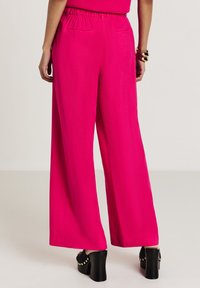 Donna che indossa pantaloni a gamba larga di un vivace rosa, tacchi a piattaforma neri con borchie dorate e bracciali dorati, in piedi su uno sfondo chiaro.