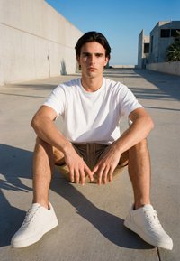 Young man in white t-shirt, beige shorts, and white sneakers sitting cross-legged on concrete outdoors with modern buildings and clear sky behind him.