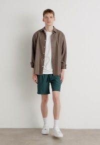 Chemise boutonnée marron clair sur un t-shirt blanc; shorts turquoise, chaussettes blanches et baskets; design minimaliste sur fond uni.