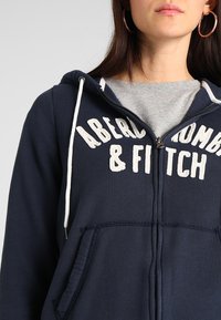 Marine blauwe hoodie met rits, gemaakt van katoen, met witte tekst "ABERCROMBIE & FITCH" op de voorkant, een capuchon met trekkoord en voorzakken.