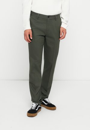 Olive green trousers with a flat front, tapered leg, and belt loops. Paired with black sneakers featuring white laces and tan soles.