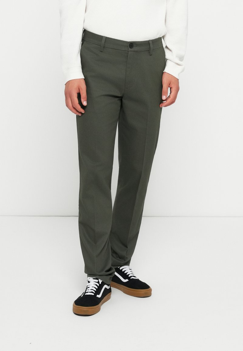 Olijfgroene broeken met een platte voorkant, taps toelopende pijpen en riemlussen. Gecombineerd met zwarte sneakers met witte veters en tan kleurige zolen.