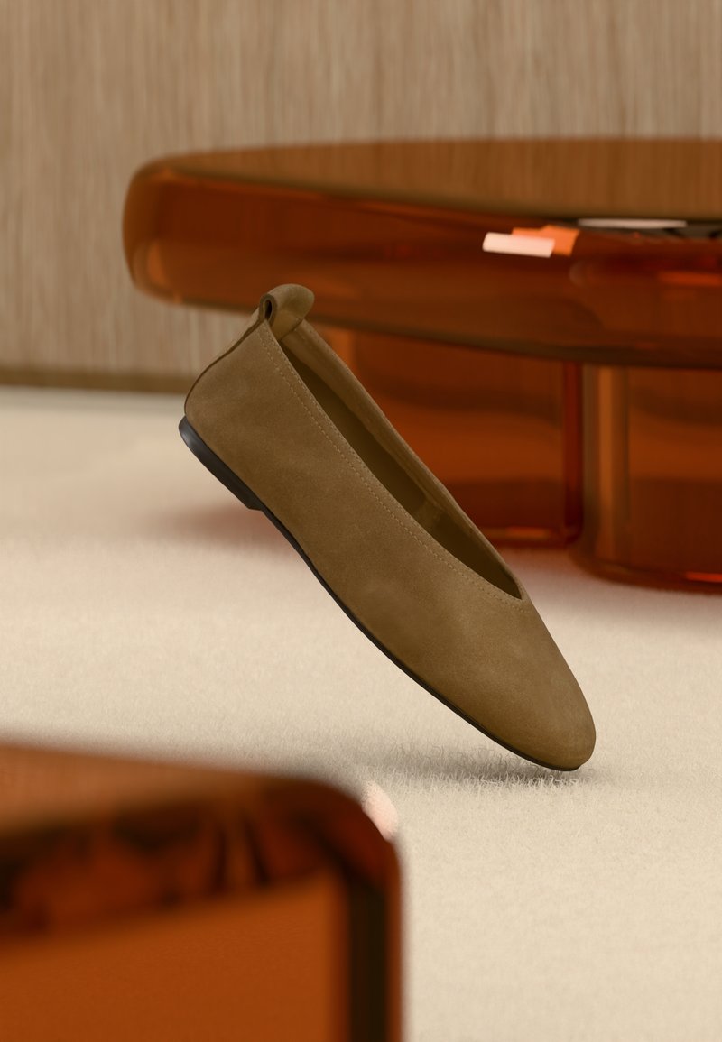 Chaussure plate en daim marron avec semelle noire flottant au-dessus d'un tapis beige, avec un mobilier translucide ambré en arrière-plan.