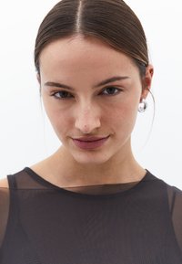 Närbild på en person som bär en genomskinlig svart topp. Har rak hår, naturlig sminkning och silverfärgade hoopsmycken. Slät hy, neutral uttryck.
