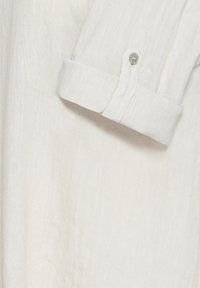 Gros plan d'une manche de chemise en lin blanc avec un revers roulé fermé par un petit bouton.