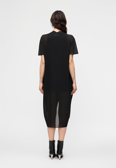 Γυναίκα με μακριά σκούρα μαλλιά, ντυμένη με διαφανές μαύρο midi φόρεμα και μαύρα μποτάκια με τακούνι, στέκεται με την πλάτη προς τα εμπρός σε μια απλή φόντο.