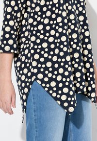 Navybluse mit cremefarbenen Punkten, mit einem dreieckigen Saum, drei Viertel-Ärmeln und einem weichen, leicht strukturierten Stoff. Getragen mit hellen Jeans.