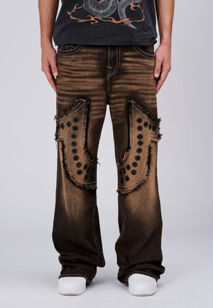 Losse bruine spijkerbroek met grote rafelige plekken en zwarte oogjes aan de voorkant van de bovenbenen, gedragen met witte sneakers en een zwart grafisch shirt.