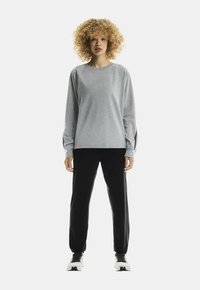 Person with curly hair stands facing forward, wearing a grey sweatshirt, black pants, and white sneakers against a plain white background.