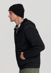 Felpa nera con zip e colletto foderato in sherpa, polsini a costine, tasche laterali, indossata sopra una camicia a quadri. Indossata con un berretto di maglia nero.