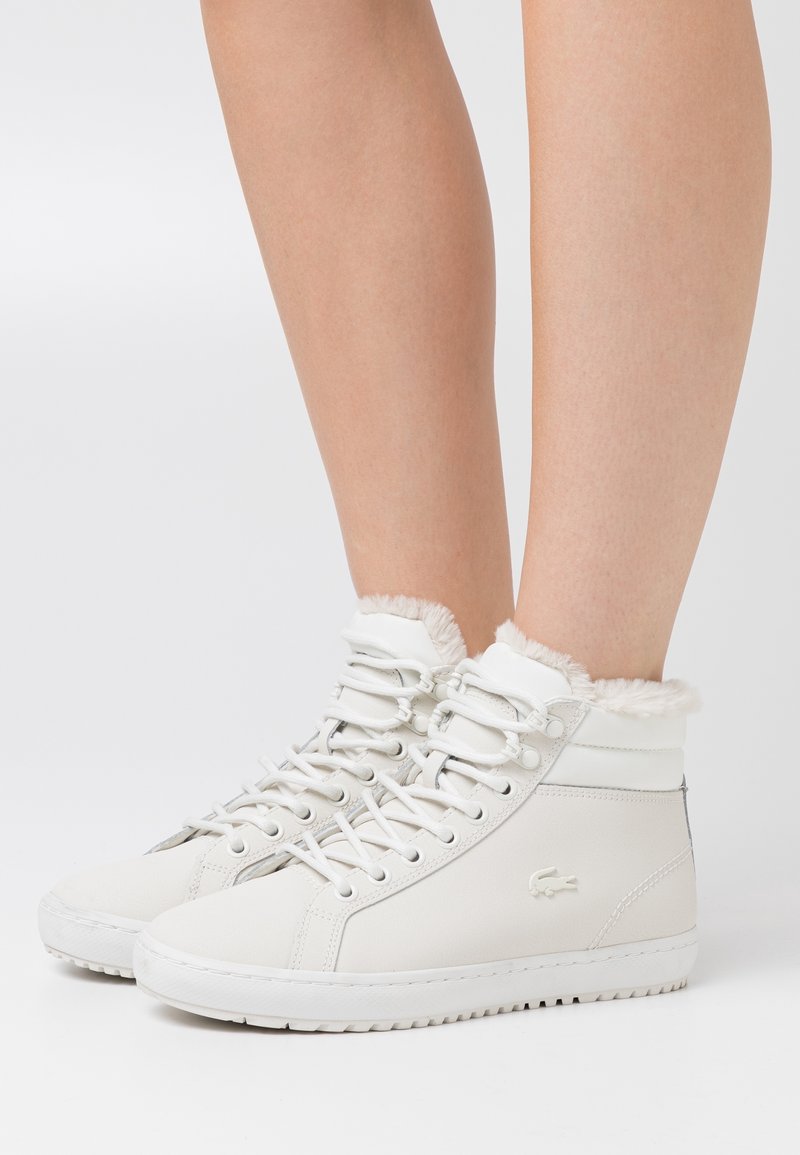 Baskets montantes blanches avec doublure en fourrure portées aux pieds, avec lacets et petit logo embossé sur le côté.