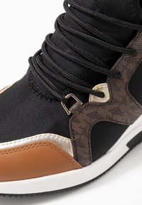 Botte de sport en maille noire avec des accents marron et or, présentant un design texturé, des lacets proéminents et une semelle en caoutchouc avec des bordures blanches.