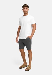 Hombre de pie vistiendo una camiseta blanca sencilla, pantalones cortos oscuros grises con bolsillos y sandalias beiges frente a un fondo claro y liso.