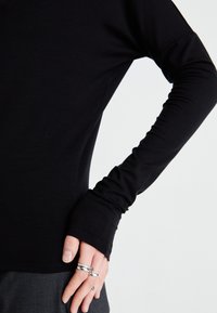 Black long-sleeve top made of smooth fabric, featuring a relaxed fit, extended cuffs, and a rounded neckline. Hand adorned with rings.