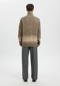 Pārmēra lieluma brūns džemperis ar ribotu augstu apkakli, kam pie apakšas ir gaisa puskoda tonēšana gaišākā bēšā krāsā. Valkāts ar pelēkām biksēm.