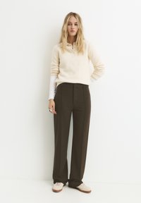 Maglione beige in maglia con colletto henley, abbinato a pantaloni a gamba larga verde scuro. La modella indossa sneakers crema e un anello argento.