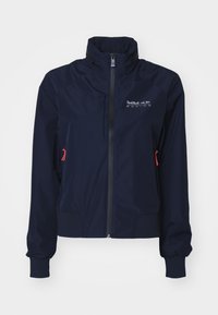 Chaqueta cortavientos azul marino con cuello alto, cremallera frontal, puños elásticos y detalles en rojo. Logo en la parte superior izquierda del pecho.