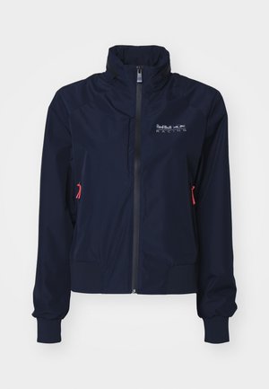 Marineblaue Windjacke mit hohem Kragen, vorderem Reißverschluss, elastischen Bündchen und roten Hardware-Details. Logo auf der oberen linken Brustseite.
