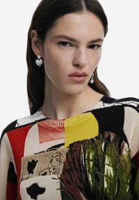 Un maglione lavorato a maglia colorato e con motivi presenta sezioni rosse, gialle e nere con design grafici. Il modello indossa orecchini a forma di cuore in argento.