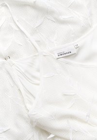 Vit plagg med en texturerad, broderad bladmönster och en mjuk inre foder. Lappen visar "Karen by Simonsen."