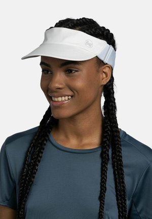 Persona sonriente con trenzas largas, que lleva una visera blanca para el sol y una camiseta deportiva azul oscuro sobre un fondo liso.