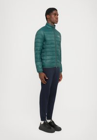 Grønn pufferjakke med høy krage og sidelukking, kombinert med marineblå tapered bukser og svarte joggesko med tykke såler.