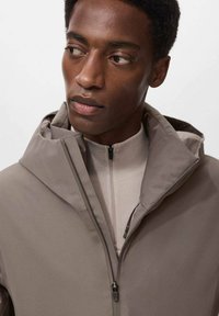Chaqueta con capucha color caqui de textura suave, con cierre de cremallera y cuello alto, combinada con una capa base de color beige claro.