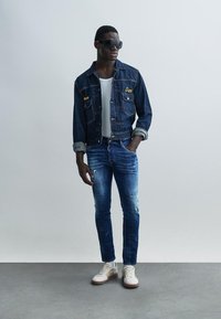 Veste en jean avec broderie jaune, débardeur blanc, jean bleu déchiré et baskets blanches avec des accents marron. Des lunettes de soleil complètent le look.