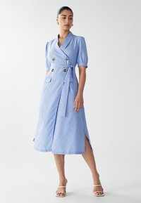 Robe portefeuille rayée bleue et blanche avec des manches courtes à volants, boutons à double boutonnage, ceinture à nouer et fentes latérales pour plus de flexibilité.