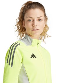 Veste de sport jaune néon avec des accents gris, fermeture éclair intégrale, col haut et trois bandes noires sur l'épaule. Présente le logo Adidas.