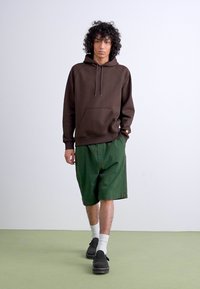 Brown, hooded sweatshirt with front pocket, paired with green shorts. Model wears black clogs and white crew socks on a light green floor.