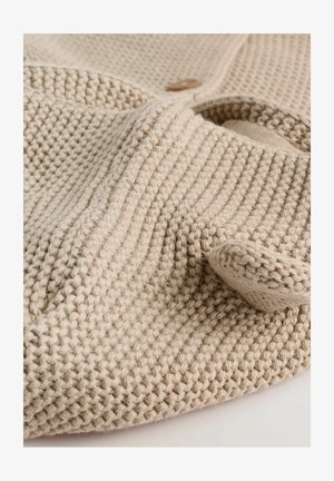 Material tricotat bej, cu un model texturat, asemănător cu un vafe. Prezintă un guler rotund și un detaliu cu un nasture din lemn. Aspect moale și confortabil.