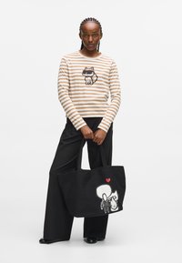 Randig långärmad tröja i beige och vitt med kattmotiv; svart tygväska med tecknad design och rött hjärta; modellen bär vida byxor.