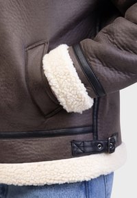 Bruine leren jas met een met schapenwol gevoerde manchette. Voorzien van zwarte leren accenten en een drukknoopsluiting op de taille, die textuurrijke materialen toont.