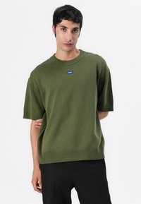 Green knit short-sleeve sweater with ribbed cuffs and hem. Features a blue logo label on the chest. Smooth texture, relaxed fit.