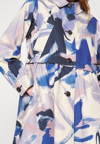 Nainen, joka pukeutuu takkiin, jossa on sinisen, mustan, vaaleanpunaisen ja valkoisen abstraktin vesivärimaalauksen kuvioita, nappikiinnitys edessä, sivutaskut ja pitkät hihat.