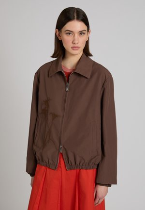 Joven con cabello castaño hasta los hombros, vistiendo una chaqueta marrón con cremallera sobre una falda roja, sobre un fondo gris y liso.