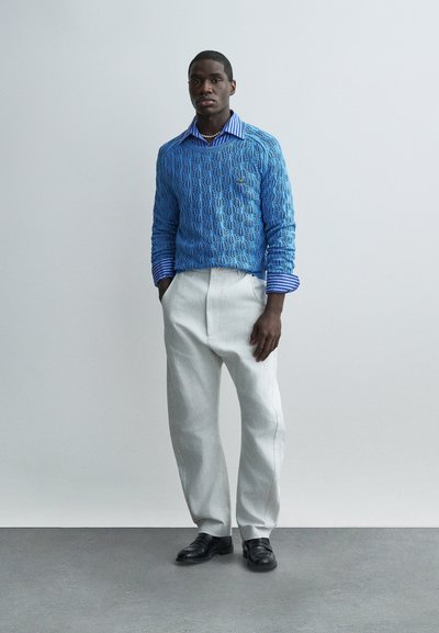 Άνδρας με μπλε πουλόβερ με σχέδια πάνω από μπλε ριγωτό πουκάμισο, ανοιχτό γκρι παντελόνι, μαύρα παπούτσια, στέκεται μπροστά σε απλό ανοιχτό γκρι φόντο.