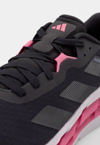 Černá sportovní bota s texturovaným svrškem, růžovými akcenty a šedými pruhy. Obsahuje ploché černé tkaničky a polstrovaný jazyk.