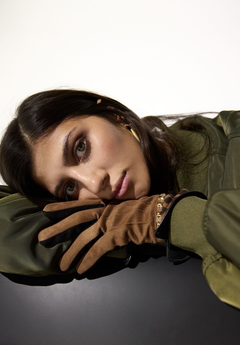 Femme aux cheveux foncés reposant la tête sur des gants en daim marron, portant une veste verte et des boucles d’oreilles dorées en anneau, sur un fond clair.
