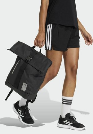 Zaino nero con chiusura a rotolo, tasca frontale, spallacci regolabili e patch con il logo. Indossato casualmente con pantaloncini neri e scarpe sportive.