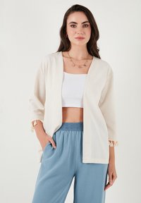 Cardigan aperto di un beige chiaro, ampio e morbido, con maniche a 3/4 e dettagli in frange, abbinato a pantaloni azzurri con coulisse e a una maglietta corta bianca.