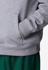 Personne portant un sweat-shirt gris clair avec une main dans la poche avant, associée à un pantalon vert foncé.