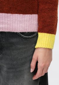 Maglione lavorato a maglia con strisce rosse, rosa e gialle e polsini a coste; abbinato a pantaloni di jeans grigi. Focus sulla texture e sul contrasto cromatico.