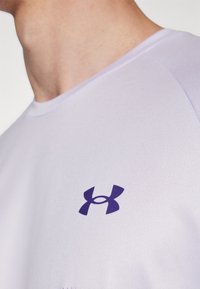 Camiseta atlética blanca con el logo morado de Under Armour en la parte izquierda del pecho. El tejido parece ligero y tiene una textura suave.