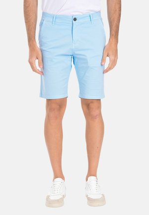 Uomo con pantaloncini azzurri al ginocchio, sneaker bianche e camicia bianca su sfondo semplice.